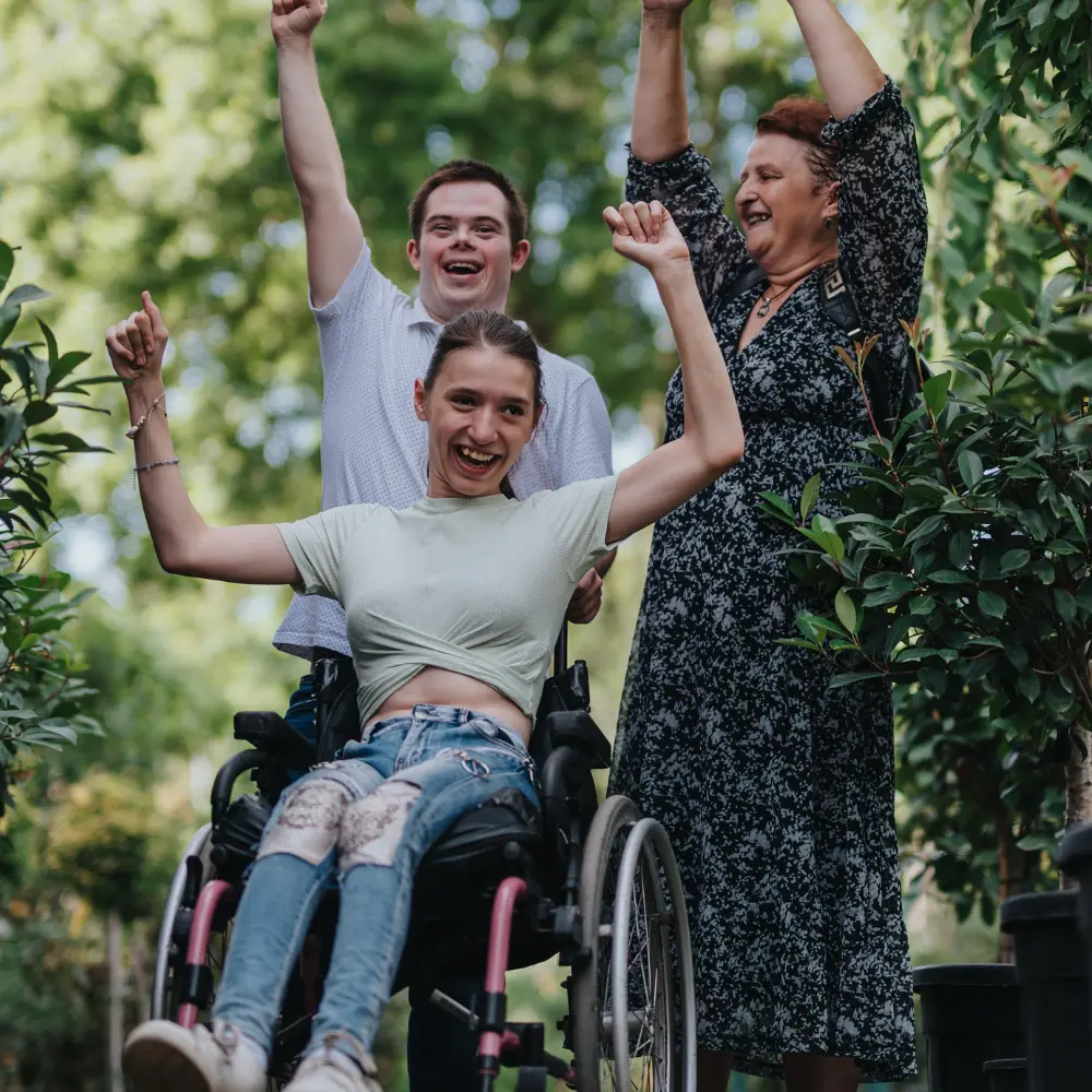 Two disabled friends with hands in the air with support worker, all smiling